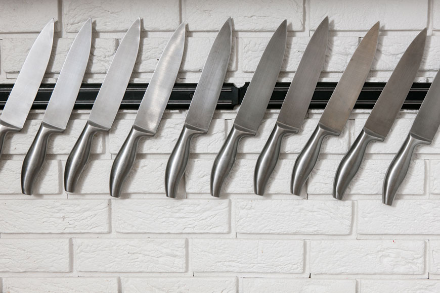 A series of chef’s knives hang on a magnetic strip on a brick wall painted white